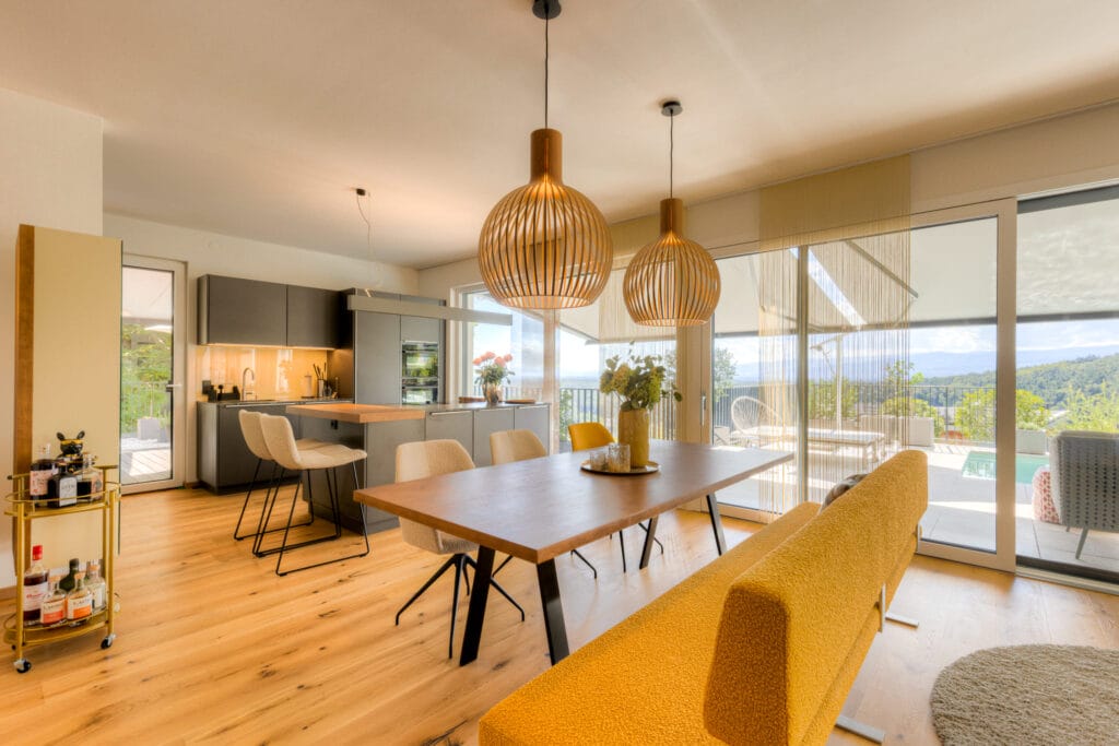 Offene Wohnküche mit Holzfußboden, grauen Küchenschränken, einem Esstisch aus Holz und einem gelben Sofa. Pendelleuchten aus Holz.
