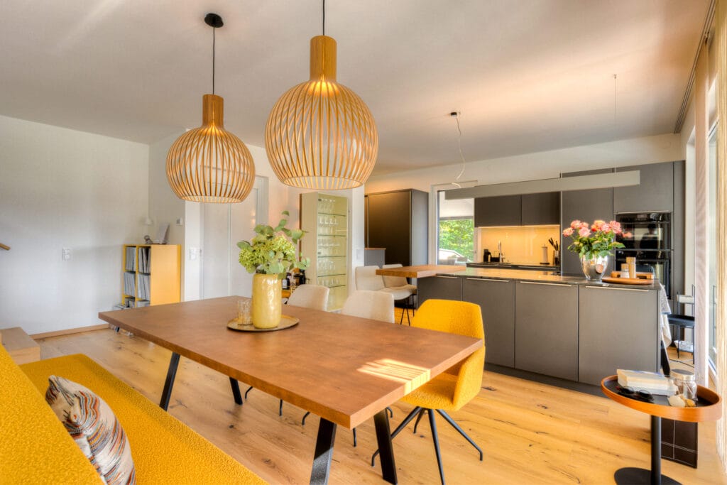 Offener Wohnraum mit Holzfußboden, grauer Küche, Essbereich mit Holztisch und orangenen Stühlen, sowie zwei geflochtenen Lampen.