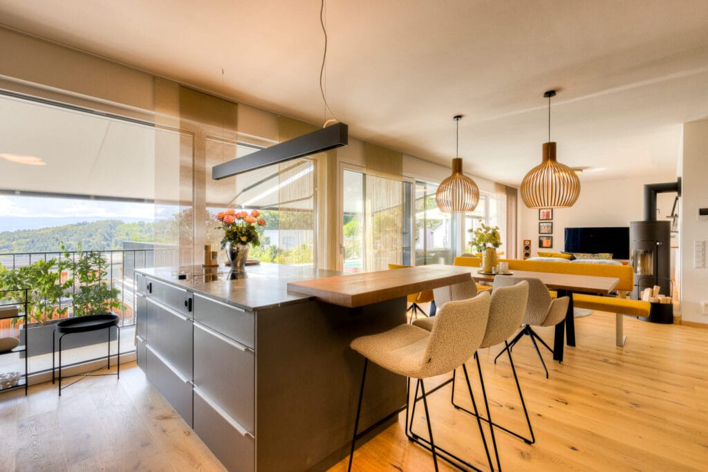 Offene Wohnküche mit Holz- und Metalloberflächen, großen Fenstern und modernen Pendelleuchten, kombiniert mit gemütlicher Sitzecke.