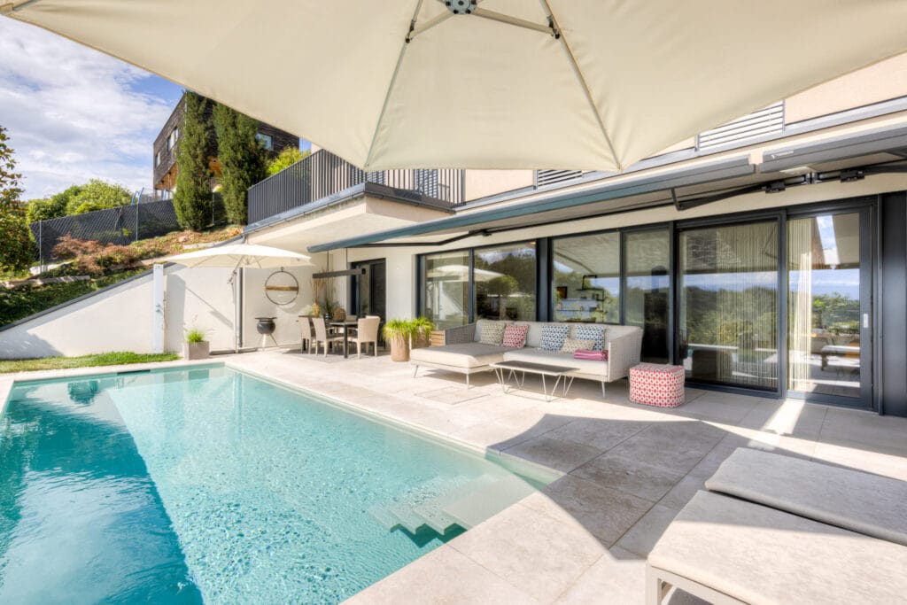 Terrasse mit Pool, Lounge-Bereich, modernen Möbeln, Fliesenboden und Blick auf Gartenlandschaft und Gartenmöbel.