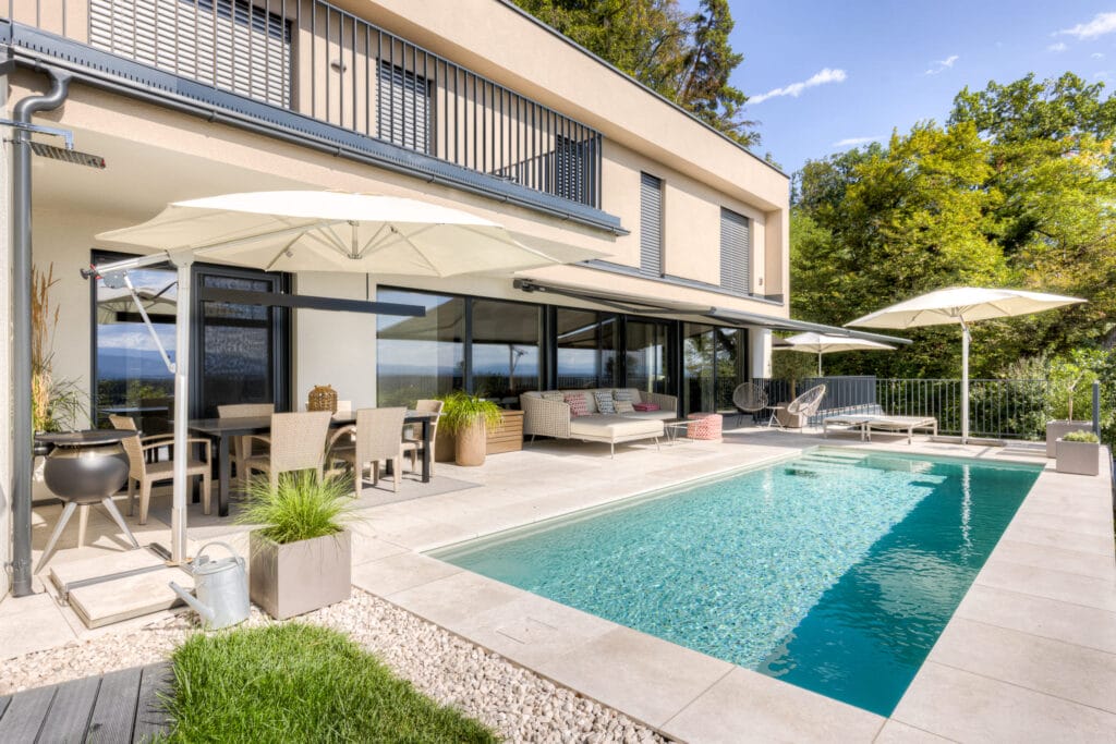 Terrasse mit Schwimmingpool, Rattanmöbeln, Sonnenschirmen und Glasschiebetüren zu einem hellen Wohnraum.