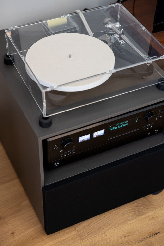 Ein Plattenspieler mit transparenter Abdeckung und eine HiFi-Anlage auf einem dunkelgrauen Möbelstück in einem hellen Raum.
