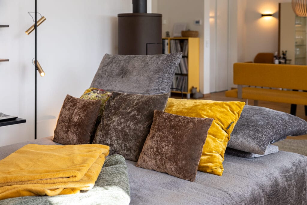 Gemütlicher Wohnraum mit einem Sofa in Grau, bunten Kissen in Gelb, Braun und Grün sowie einer Stehlampe und holzverkleideter Wand.
