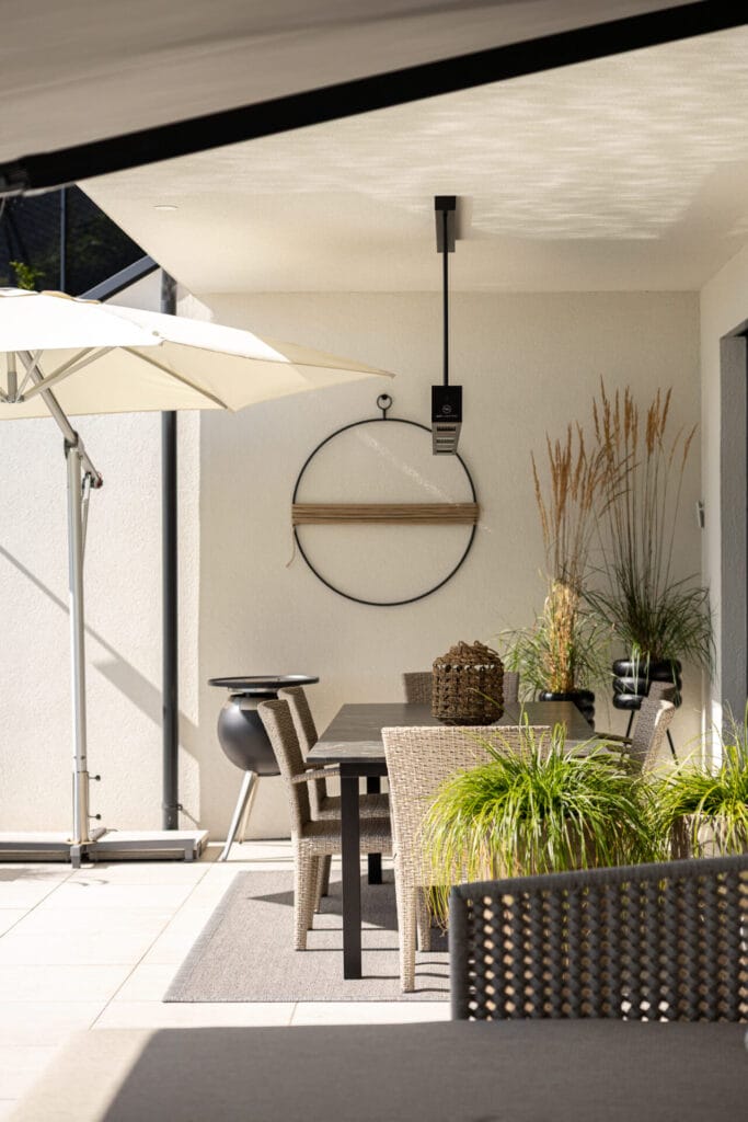 Überdachte Terrasse mit einem Essbereich, Holztisch, geflochtenen Stühlen, Pflanzen und Sonnenschirm auf hellem Fliesenboden.