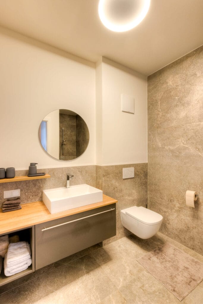 Badezimmer mit grauen Natursteinfliesen, Holzwaschbecken, runder Spiegel und minimalistischem Wandregal.