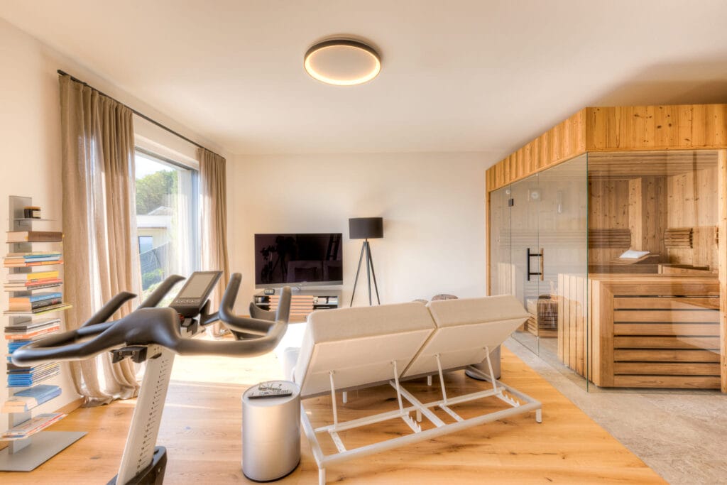 Wohnraum mit Holzfußboden, Fitnessgerät, Sofa, Fernseher und gläserner Sauna, umgeben von beige Vorhängen und Holzdetails.