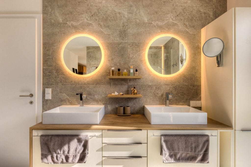 Badezimmer mit zwei weißen Waschbecken, Holzregal, großen runden, beleuchteten Spiegeln und grauen Fliesen an der Wand.