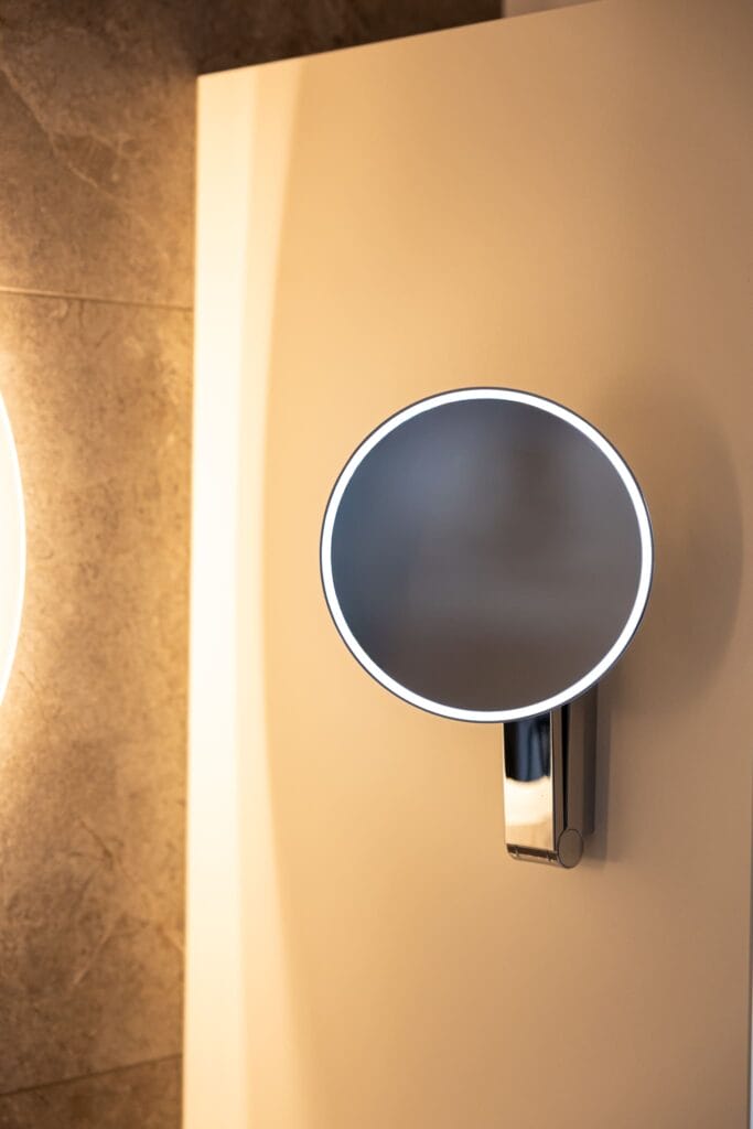 Ein runder Kosmetikspiegel mit Beleuchtung an einer hellen Wand, neben einer grauen Fliesenfläche im modernen Badezimmer.