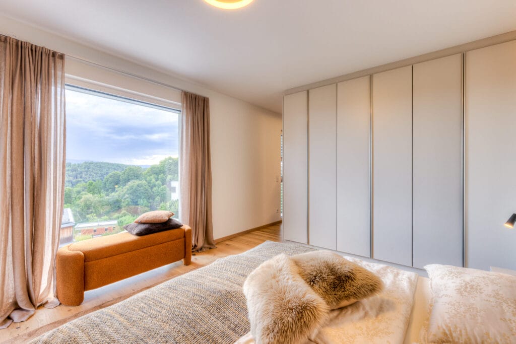 Schlafzimmer mit großen Fenstern, hellen Vorhängen, einem orangefarbenen Sofa und einem Kleiderschrank in neutralen Farben.