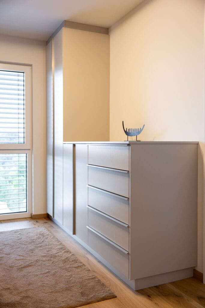 Moderner Wohnraum mit grauem sideboard, natürlichem Holzfußboden und Fenster mit Jalousien. Beige Wände und Teppich.