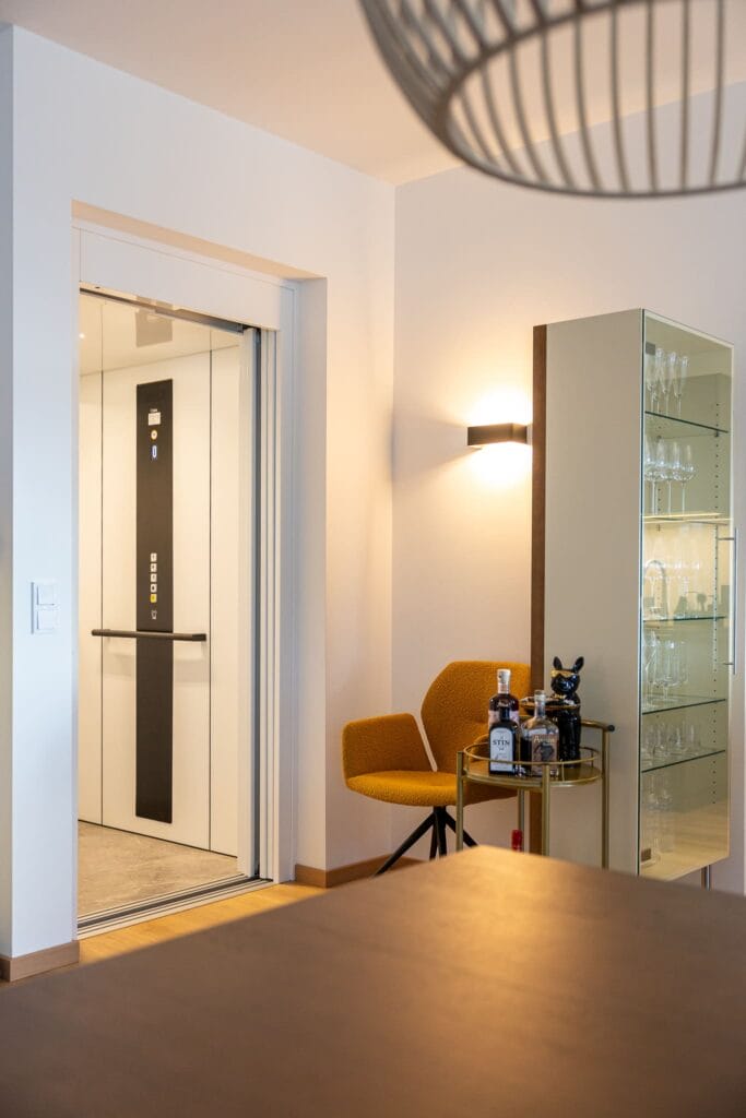 Moderne Lobby mit gläsernem Aufzug, einem orangefarbenen Sessel und einem Glastisch für Getränke, helles Wanddesign.