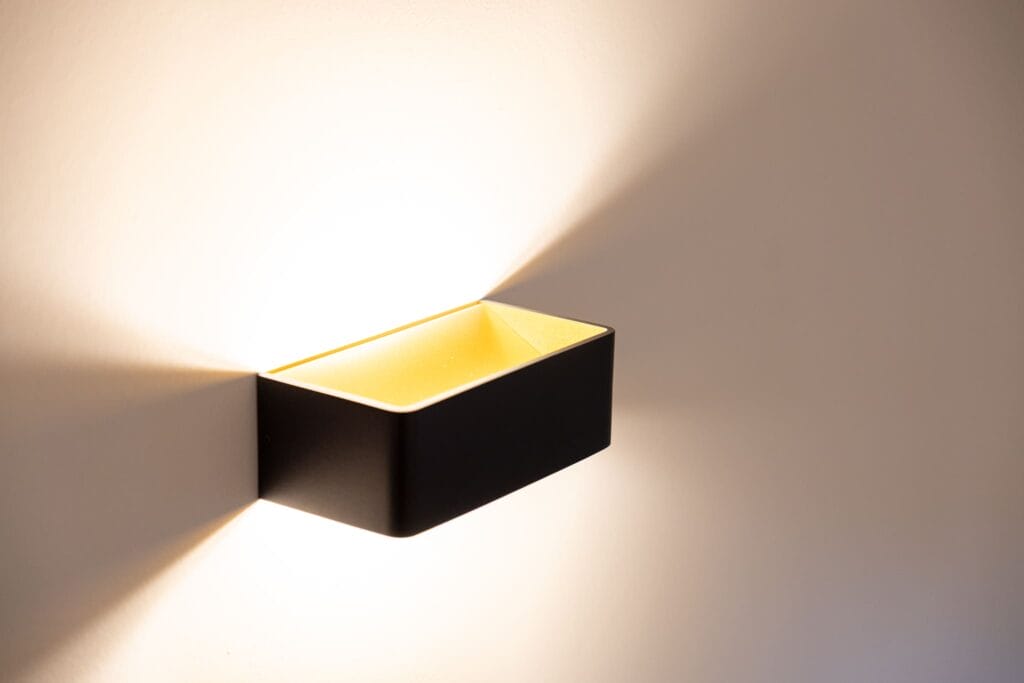 Wandleuchte in rechteckiger Form aus schwarzem Material, LED-Beleuchtung sorgt für warmes Licht auf einer weißen Wand.