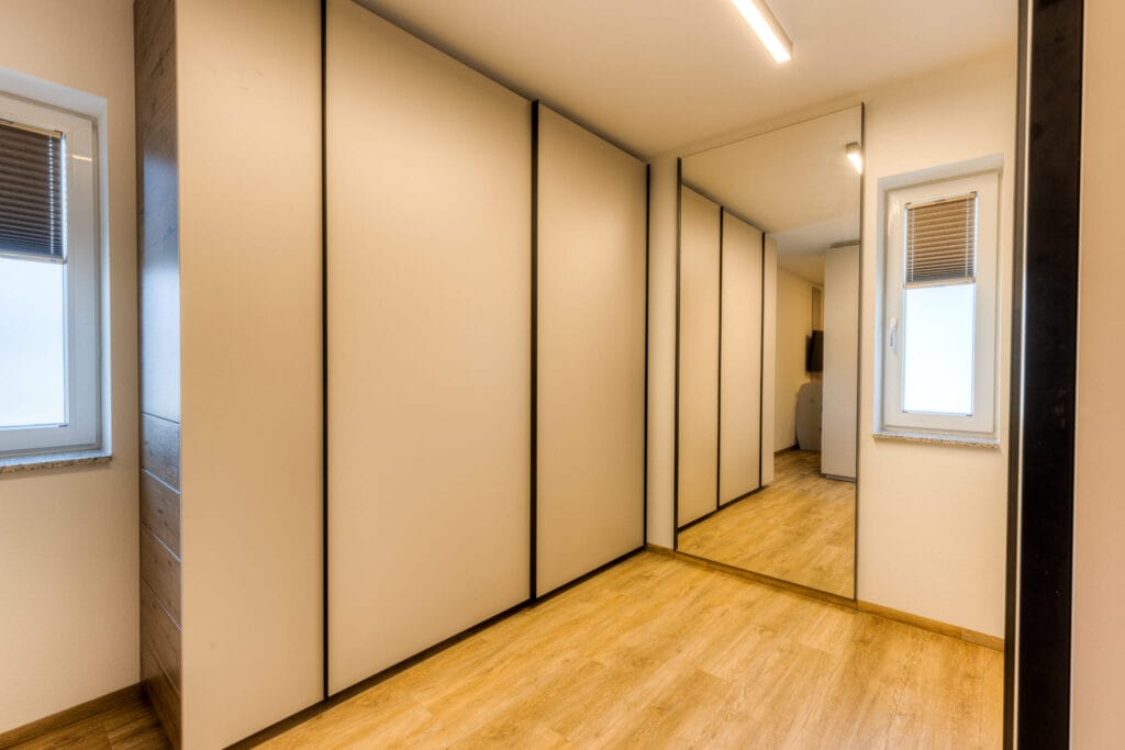 Begehbarer Kleiderschrank mit hellem Laminatboden, Wandspiegel und integrierten Schranktüren in neutralen Farben.