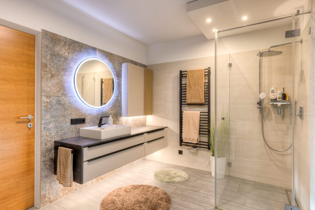 Ein modernes Badezimmer mit einer Glaskabine, einer Wandspiegelbeleuchtung, Holz- und Keramikelementen in neutralen Tönen.