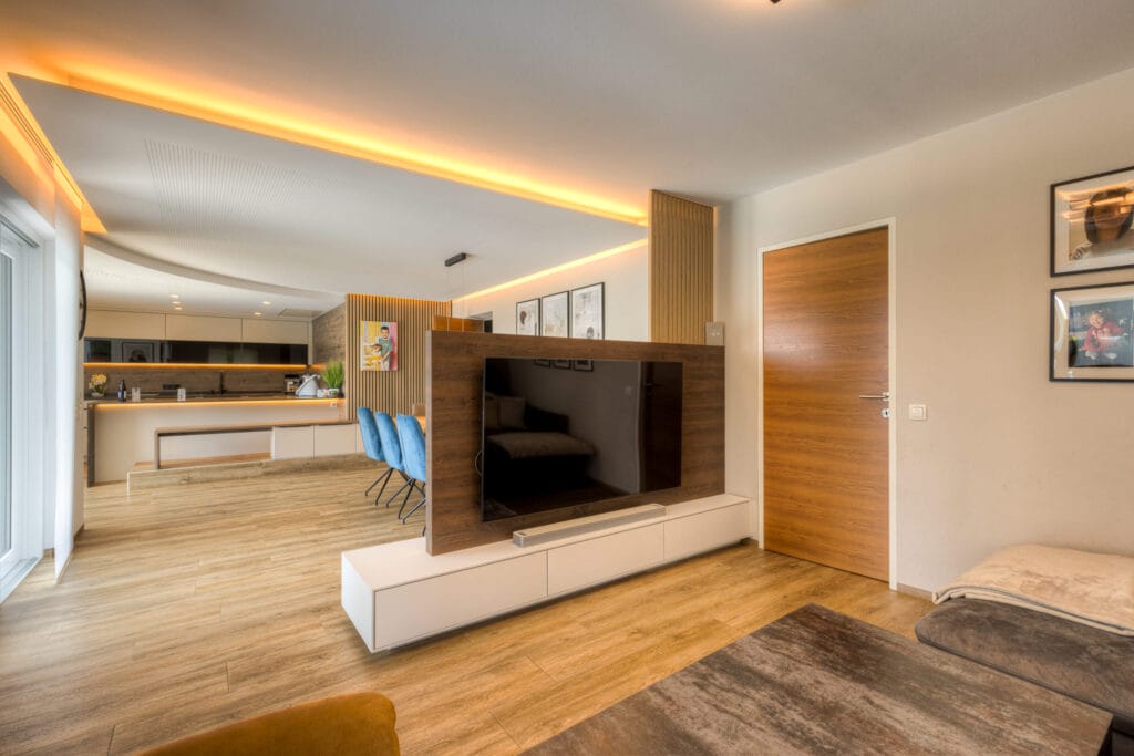 Offener Wohnraum mit Küche, Holzfußboden, fließendem Licht und einer Trennwand mit integriertem Fernseher.