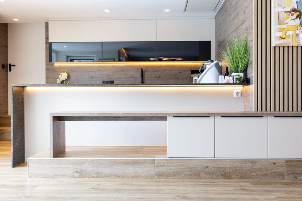 Moderne Küche mit Holz- und Granitoberflächen, Einbauküchenschränken in hellen Tönen und LED-Beleuchtung. Eine Sitzbank entlang der Kücheninsel mit indirekter Beleuchtung