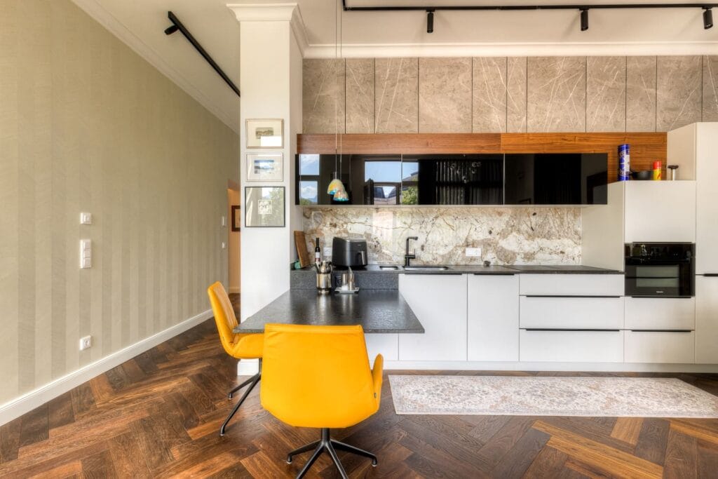 Moderne Küche mit Hochschrank, hellen Holzfronten, Marmorrückwand und orangefarbenem Stuhl auf Fischgrät-Parkettboden.