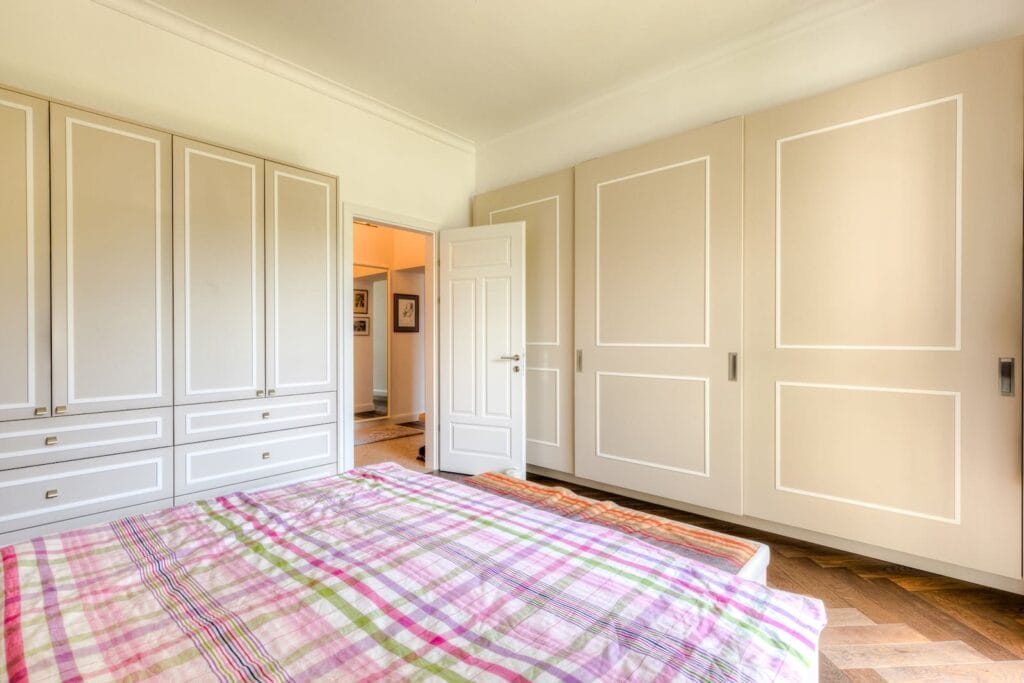 Schlafzimmer mit einem großen, beigen Schrank, Holzboden und gestreiftem Bettzeug in sanften Farben.