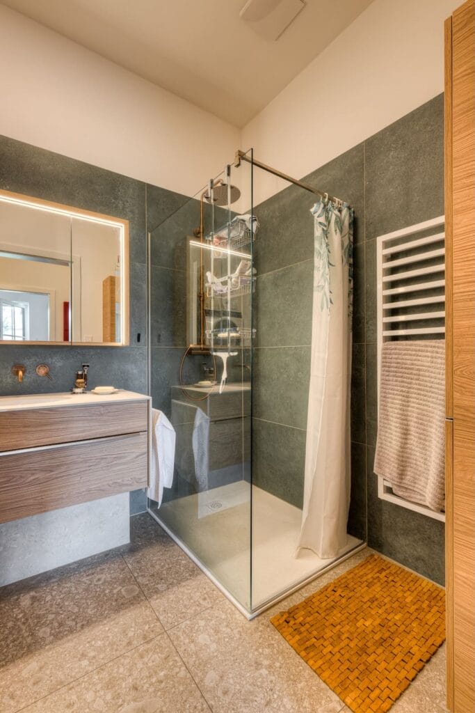 Badezimmer mit bodengleicher Dusche, Holzwaschtisch, fliesenverkleideten Wänden in grün-grauen und beige Tönen sowie Holzdetails.